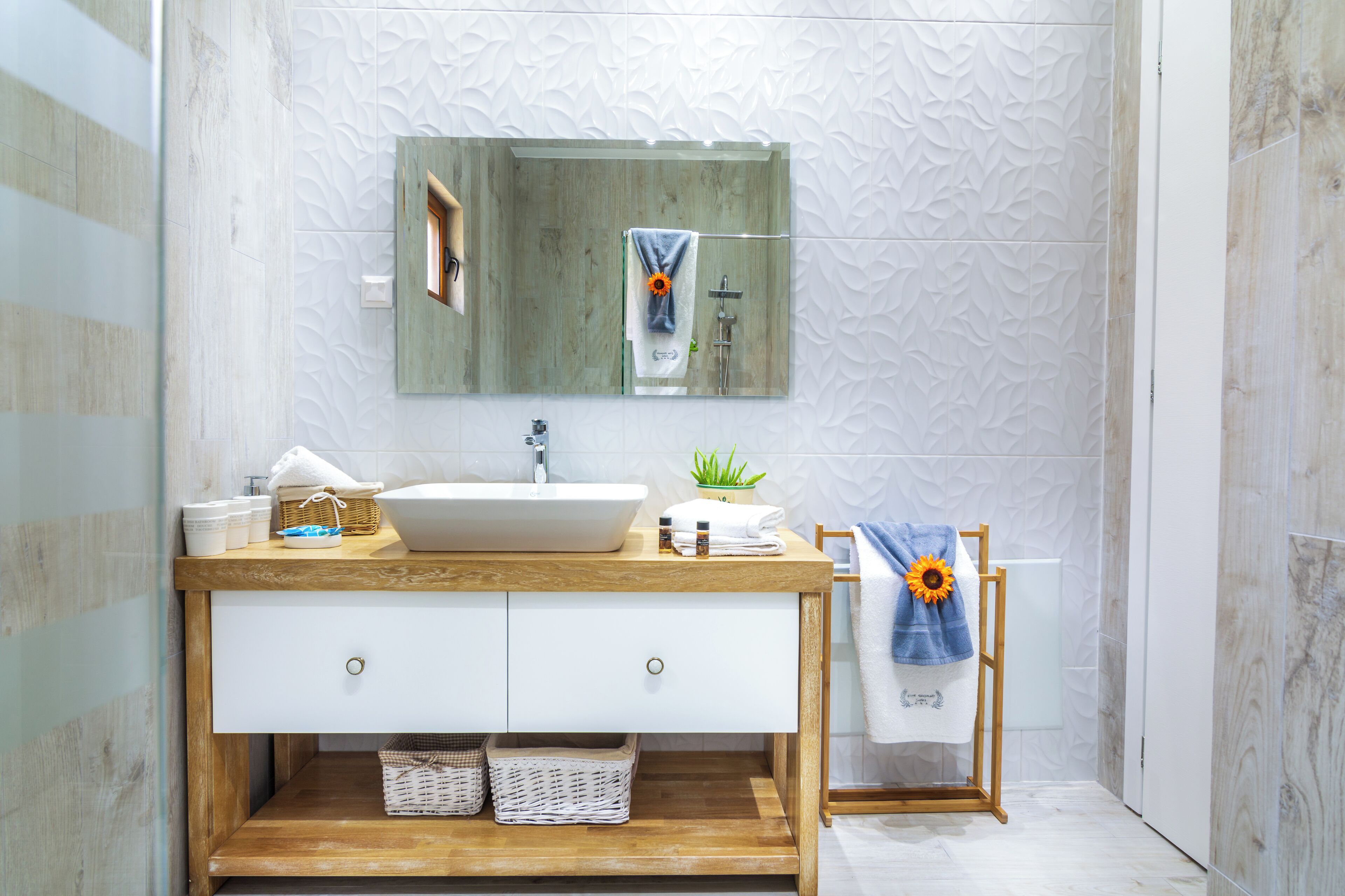 A modern bathroom with a wooden vanity, white sink, and a large mirror. There is a towel rack with a towel and a sunflower decoration.