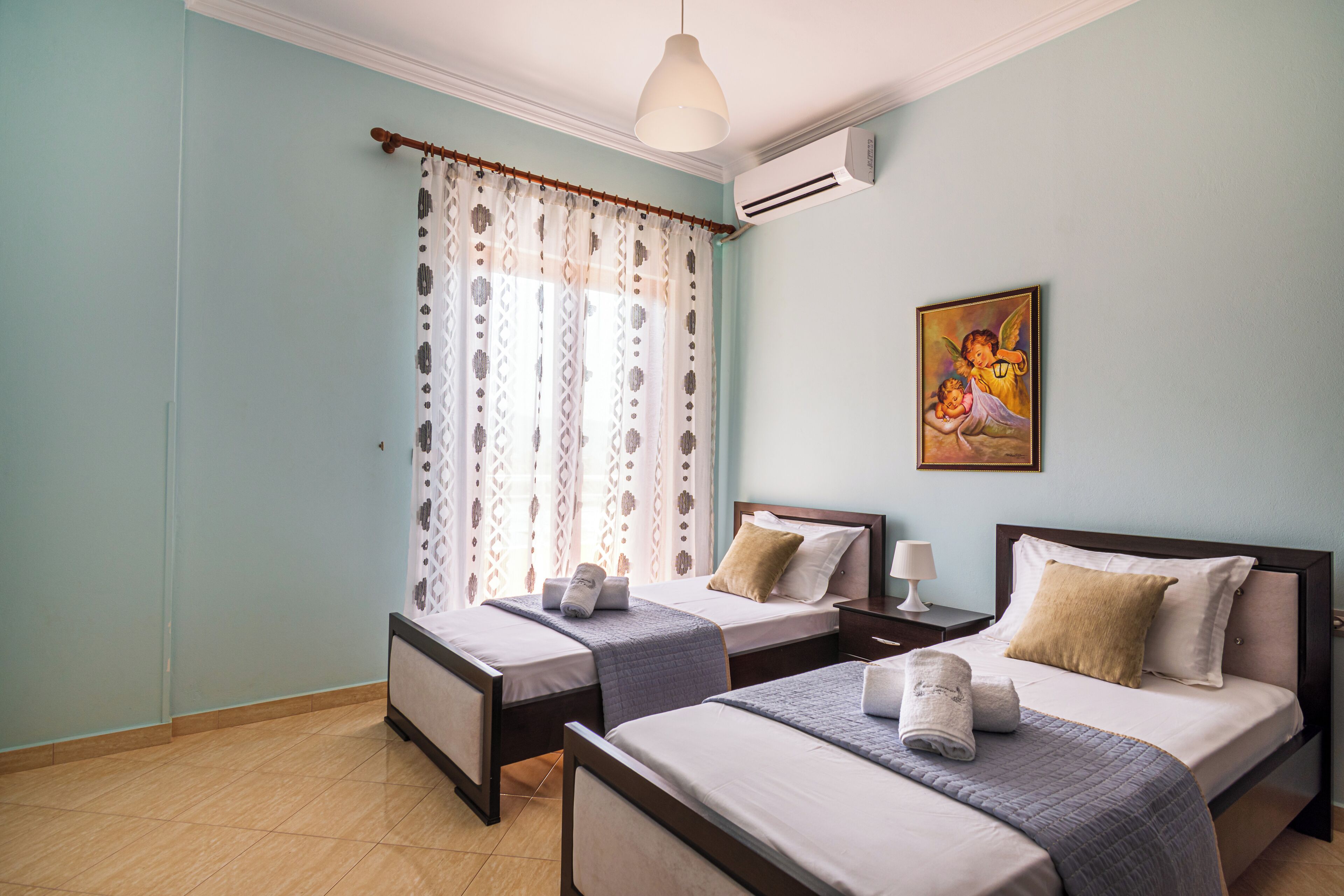 A hotel room with two single beds, a large window with patterned curtains, an air conditioning unit, and a painting on the wall.