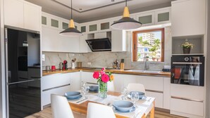 A modern kitchen with white cabinetry, a dining table set for four, and a window overlooking a residential area. - Great Alexander Suites (Sarande)