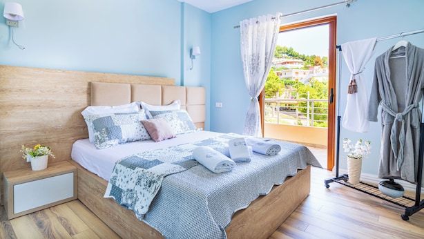 A bedroom with a wooden headboard, a bed with blue and white bedding, a nightstand with a plant, and a view of a balcony with a railing. - Great Alexander Suites (Sarande)