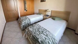 A hotel room with two beds, a wooden wardrobe, a nightstand with a lamp, and two wall-mounted paintings.