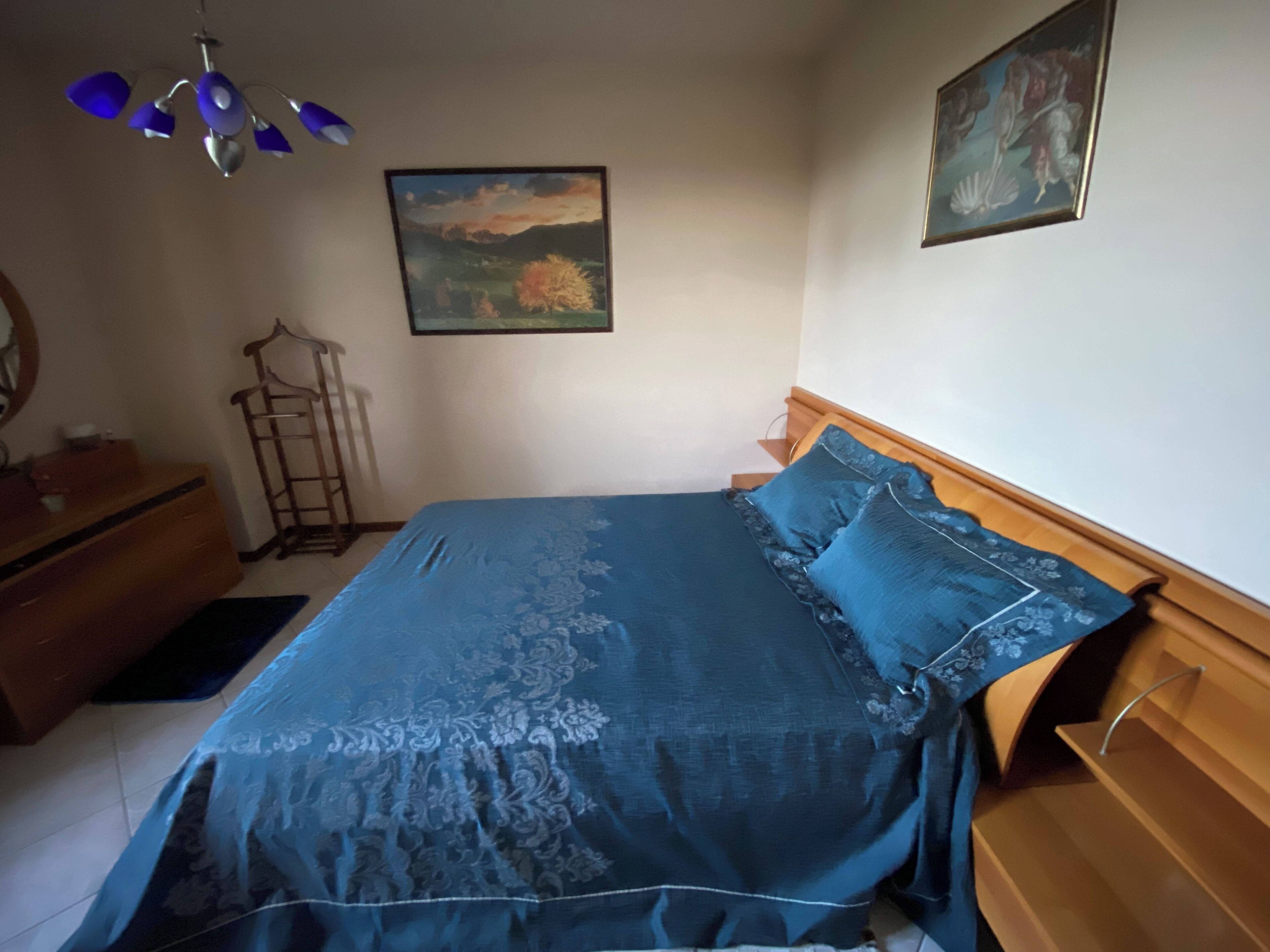 Ein Schlafzimmer mit einem Bett, einem hölzernen Kopfteil, einer Kommode, einem Spiegel, einem Kleiderständer und zwei gerahmten Bildern an der Wand.