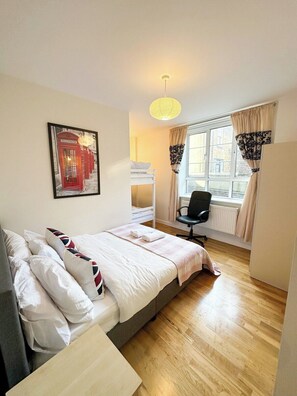 Ein Schlafzimmer mit Bett, Schreibtisch, Stuhl, einem Fenster mit Vorhängen, einem Heizkörper und einer Wandleuchte.