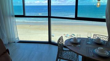 Quarto com uma janela grande dando para uma praia, mesa de jantar posta com pratos e copos, e uma cadeira.