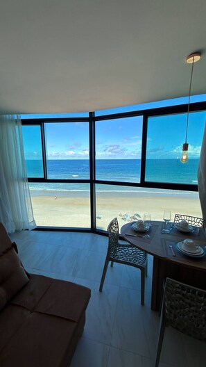 Ein Zimmer mit großem Fenster mit Blick auf einen Strand, einem Esstisch gedeckt mit Tellern und Gläsern sowie einem Stuhl.
