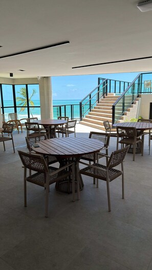 Un'ampia area interna con diversi tavoli in legno e sedie in vimini, una scala che conduce a un balcone esterno e vista sull'oceano.