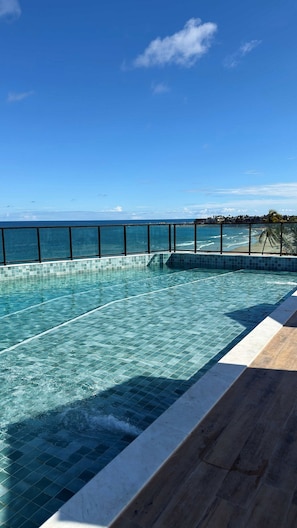 A swimming pool with clear blue water, a wooden deck, and a view of the ocean and beach. - Vila Porto Beira Mar (Ipojuca)