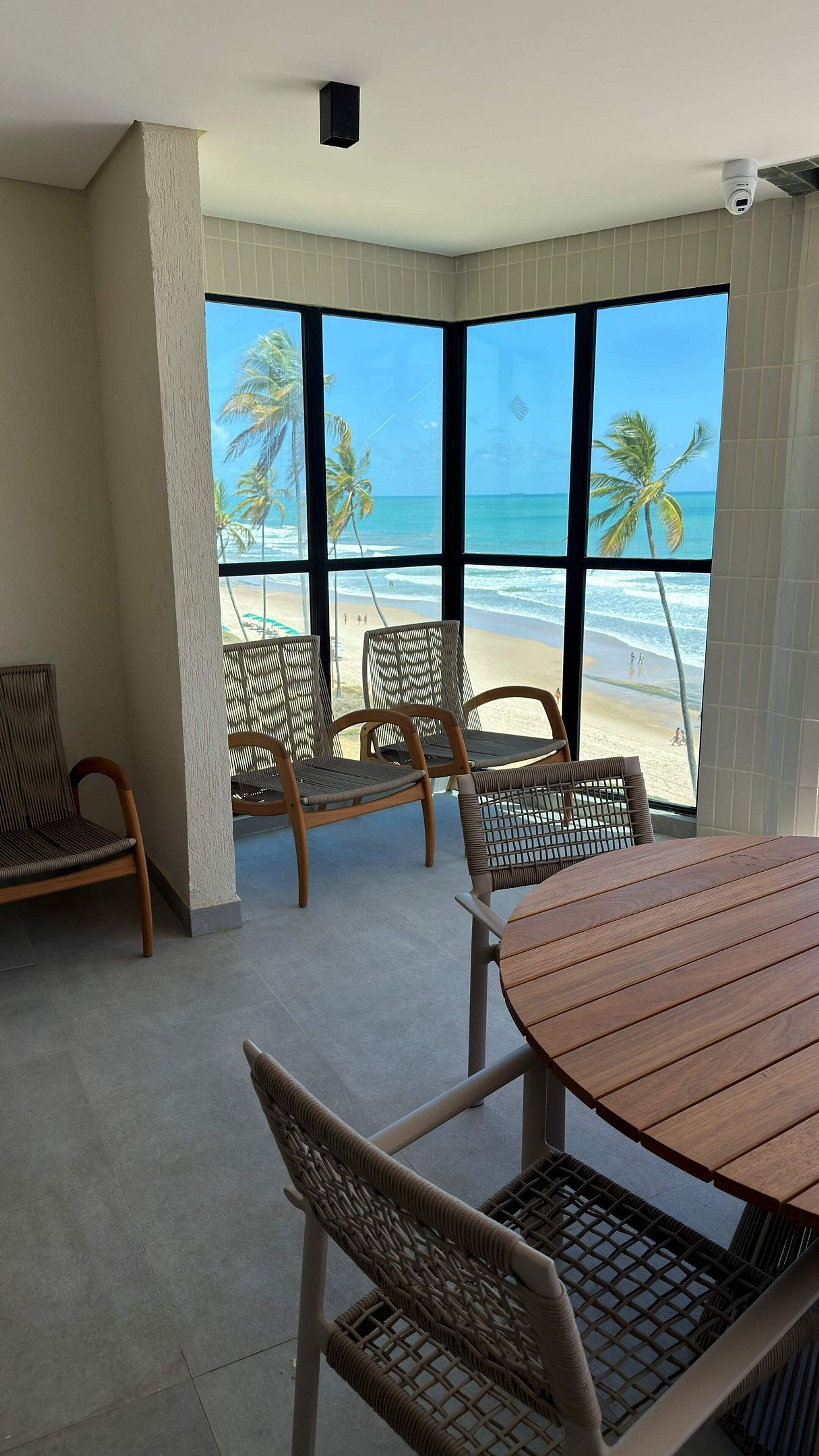 Quarto com uma grande janela com vista para a praia, mobiliário de vime e uma mesa redonda de madeira.