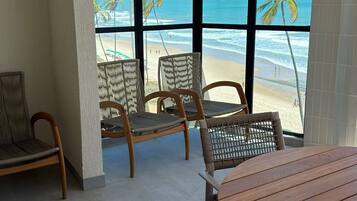 Quarto com uma grande janela com vista para a praia, mobiliário de vime e uma mesa redonda de madeira.