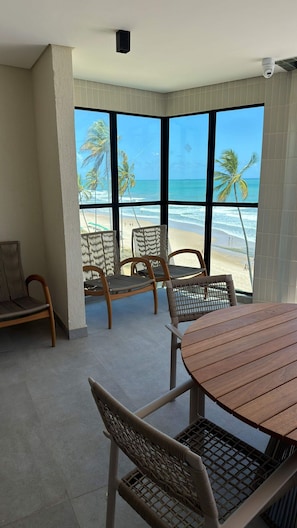 A room with a large window overlooking a beach, wicker furniture, and a round wooden table. - Vila Porto Beira Mar (Ipojuca)