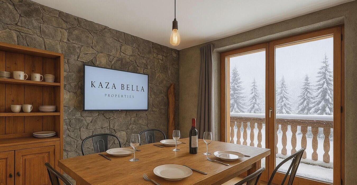 Zona de menjador amb una paret de pedra, un cartell emmarcat que diu "KAZA BELLA PROPERTIES", una taula de fusta preparada per a quatre i vista a arbres coberts de neu a través d'una porta de balcó.