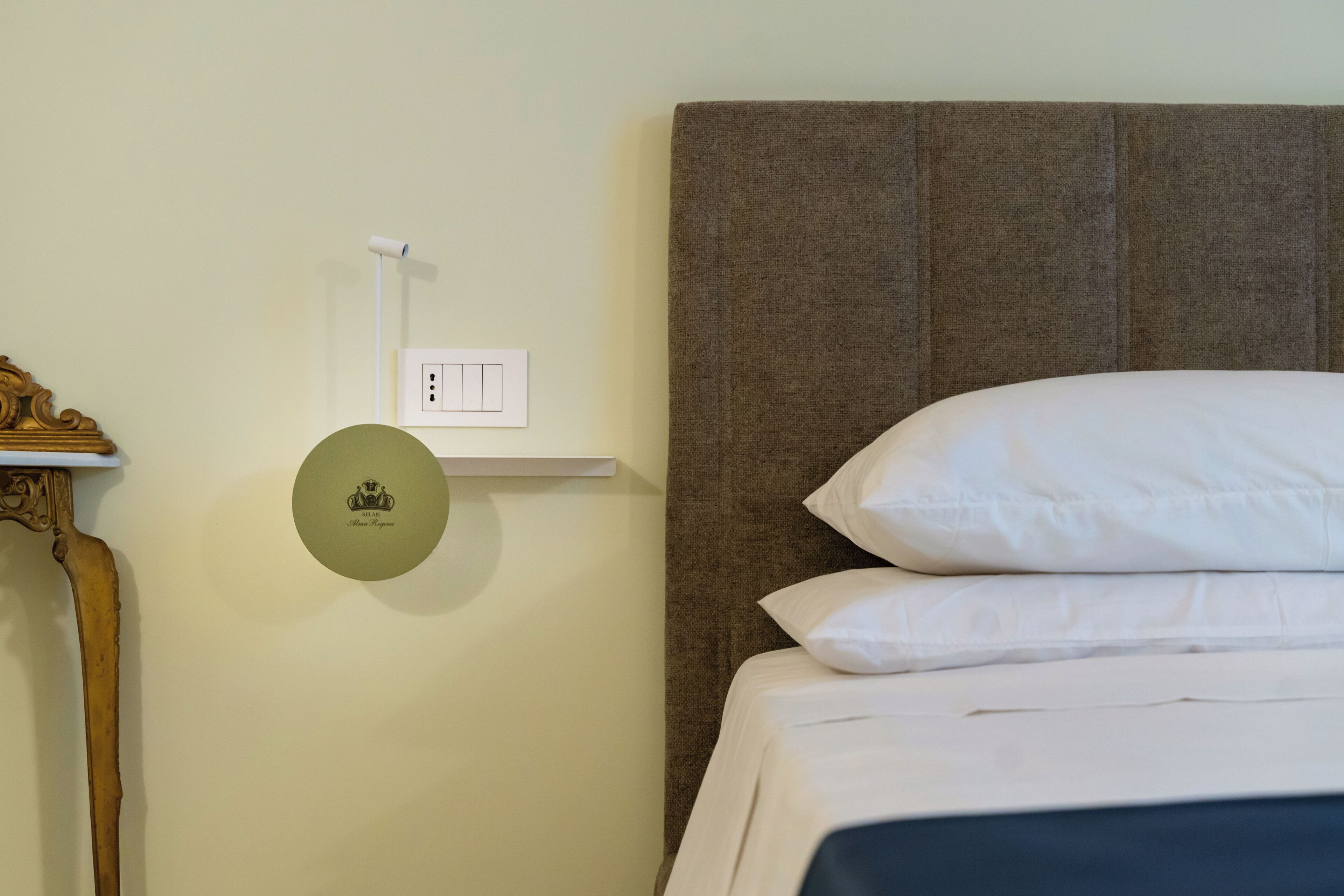 A neatly made bed with white linens and a brown headboard, a wall-mounted power outlet, and a small shelf with a decorative item.