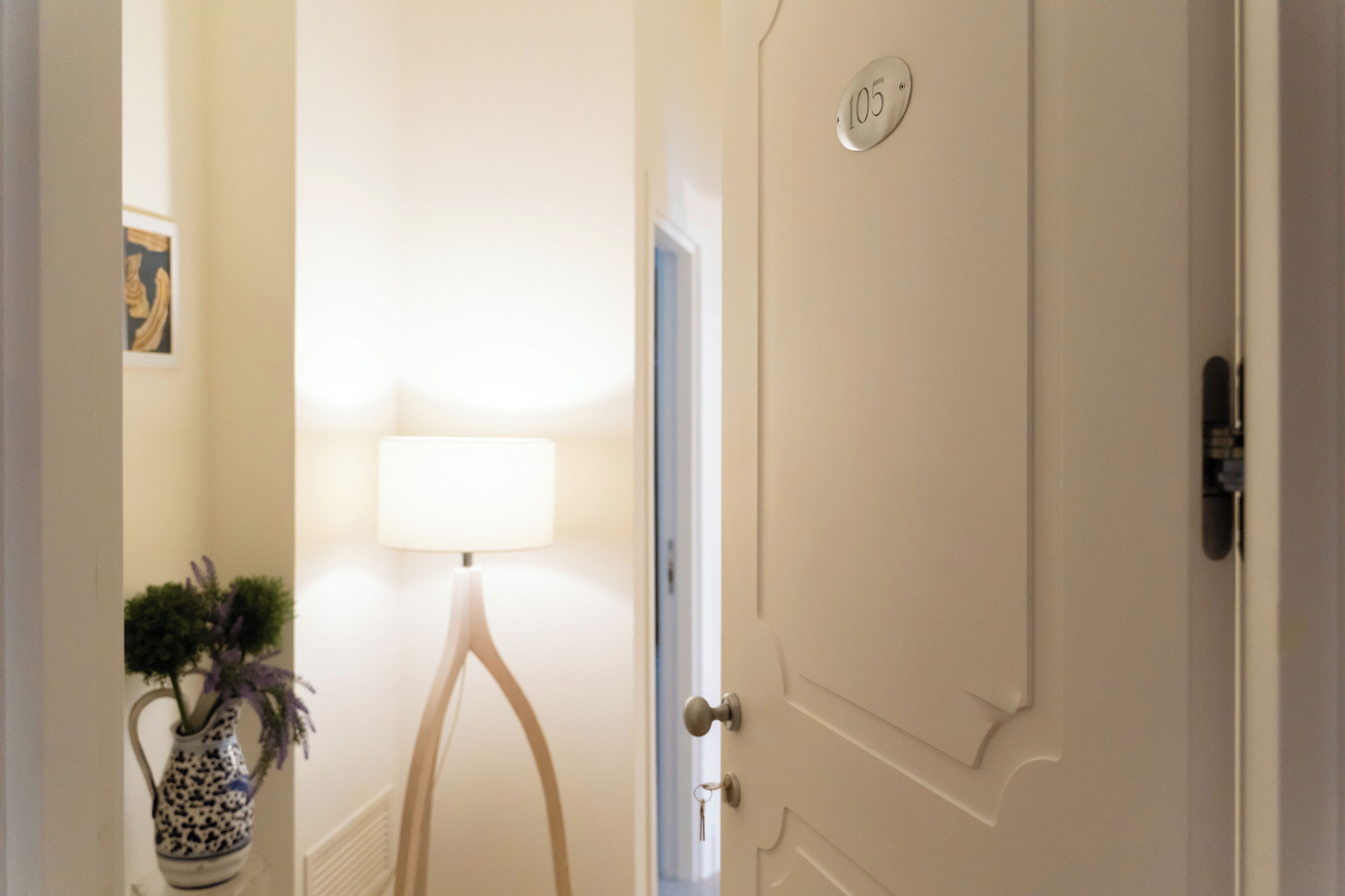 Una habitación de hotel con la puerta cerrada, una lámpara y un florero con flores.