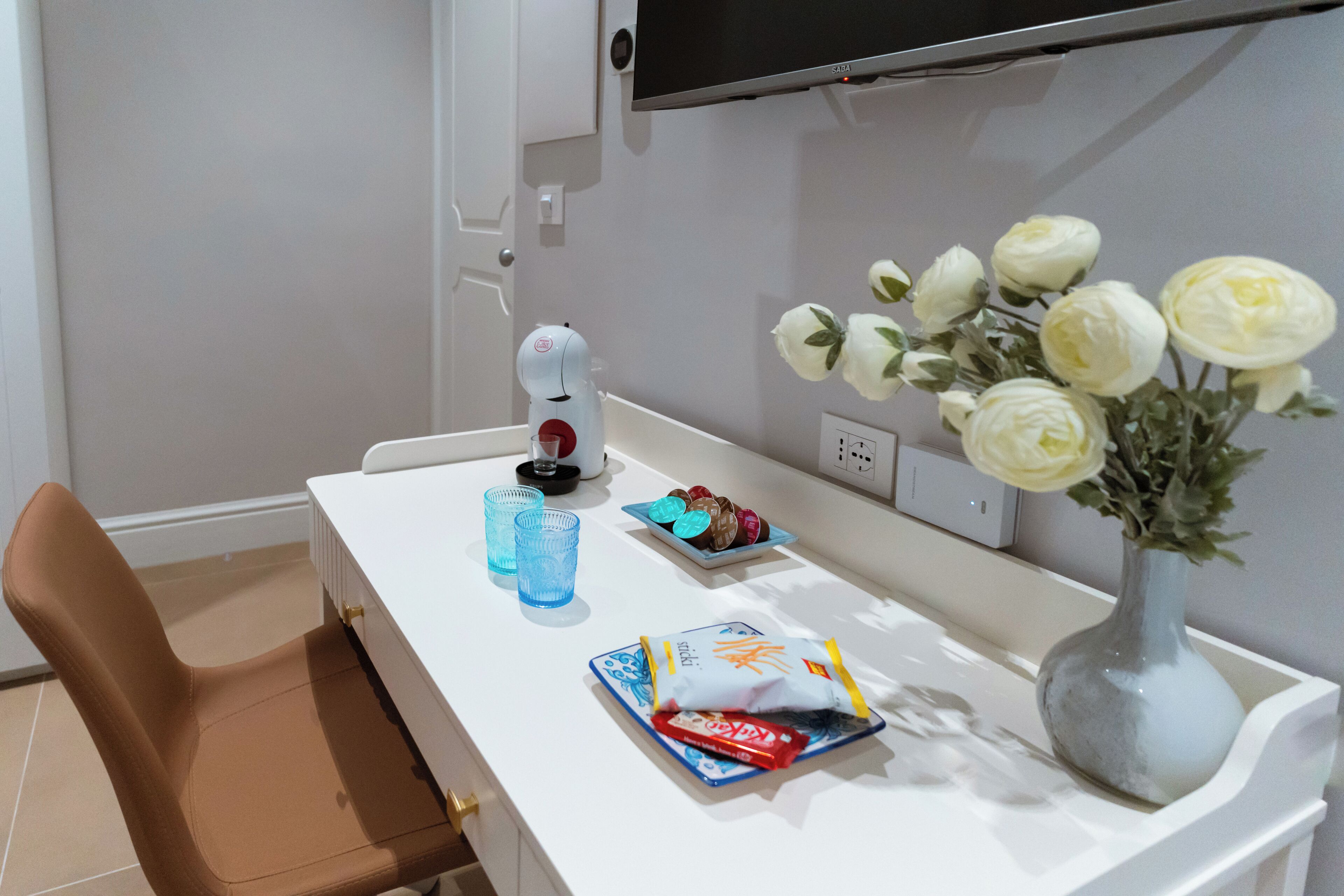 A hotel room with a white desk, a vase with white flowers, a blue glass, a plate with snacks, and a bag of chips.