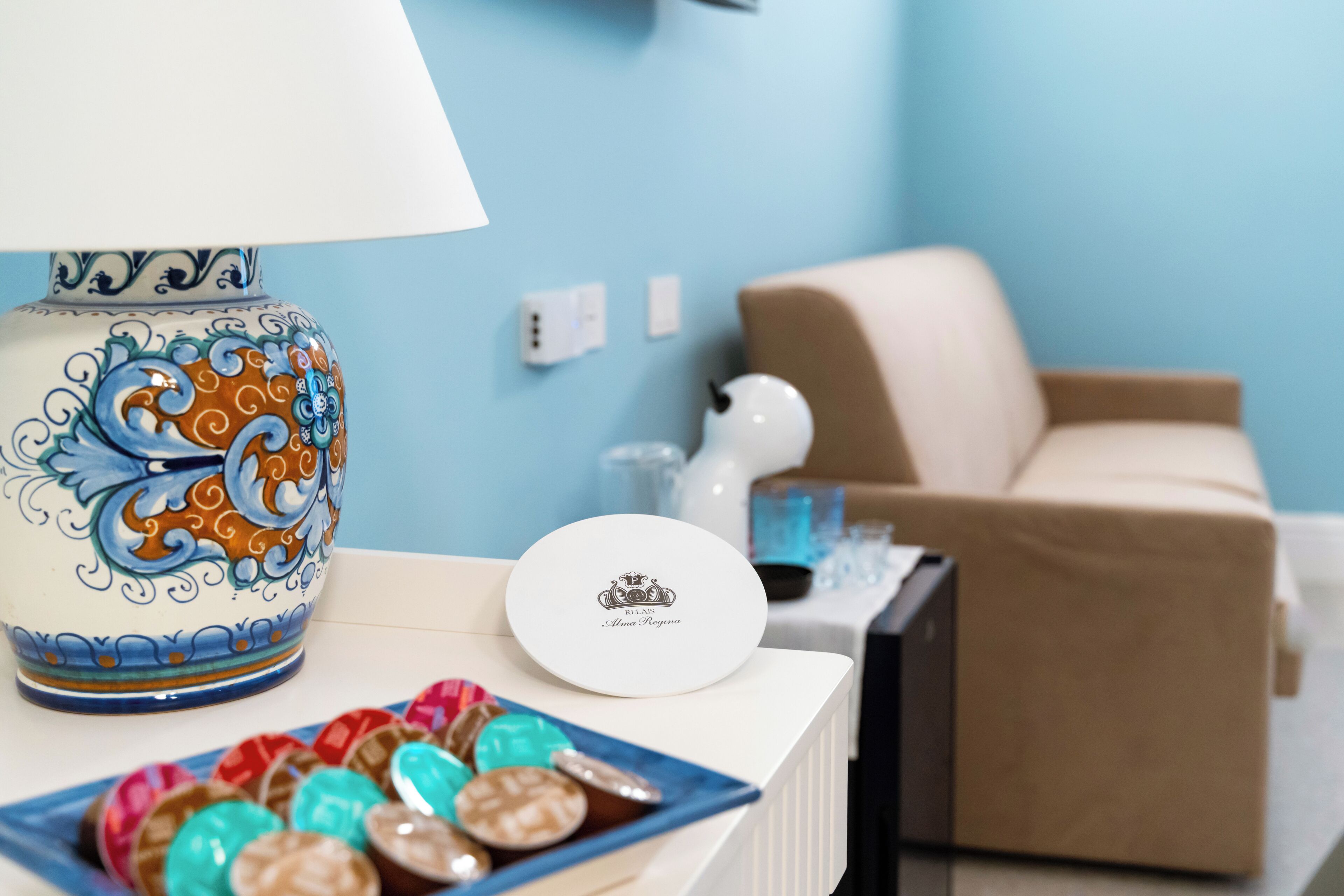 A decorative ceramic vase, a white plate with a logo, and a plate of colorful chocolates on a table in a room with a blue wall and a beige sofa.