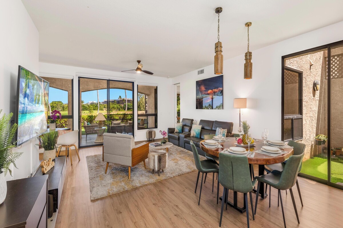 A spacious living area with a dining table set for four, a sofa, and a large window offering a view of the outdoors.