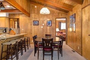 Una habitación con paneles de madera, una mesa redonda para comer, varias sillas, una zona de cocina con cafetera y cuadros en las paredes.