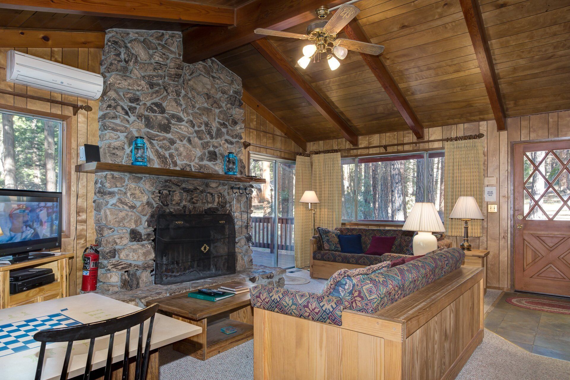 A cozy cabin interior with a stone fireplace, wooden furniture, a television, and a ceiling fan.
