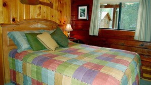 Ein Schlafzimmer einer Holzhütte mit einem Bett, einem Nachttisch, einer Lampe, einem Fenster mit Vorhängen und einer Holzwand, an der eine Gitarre befestigt ist.