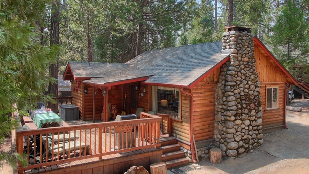 A log cabin with a stone chimney, wooden deck, and a covered outdoor seating area. - Chipmunks Holiday (Yosemite National Park)