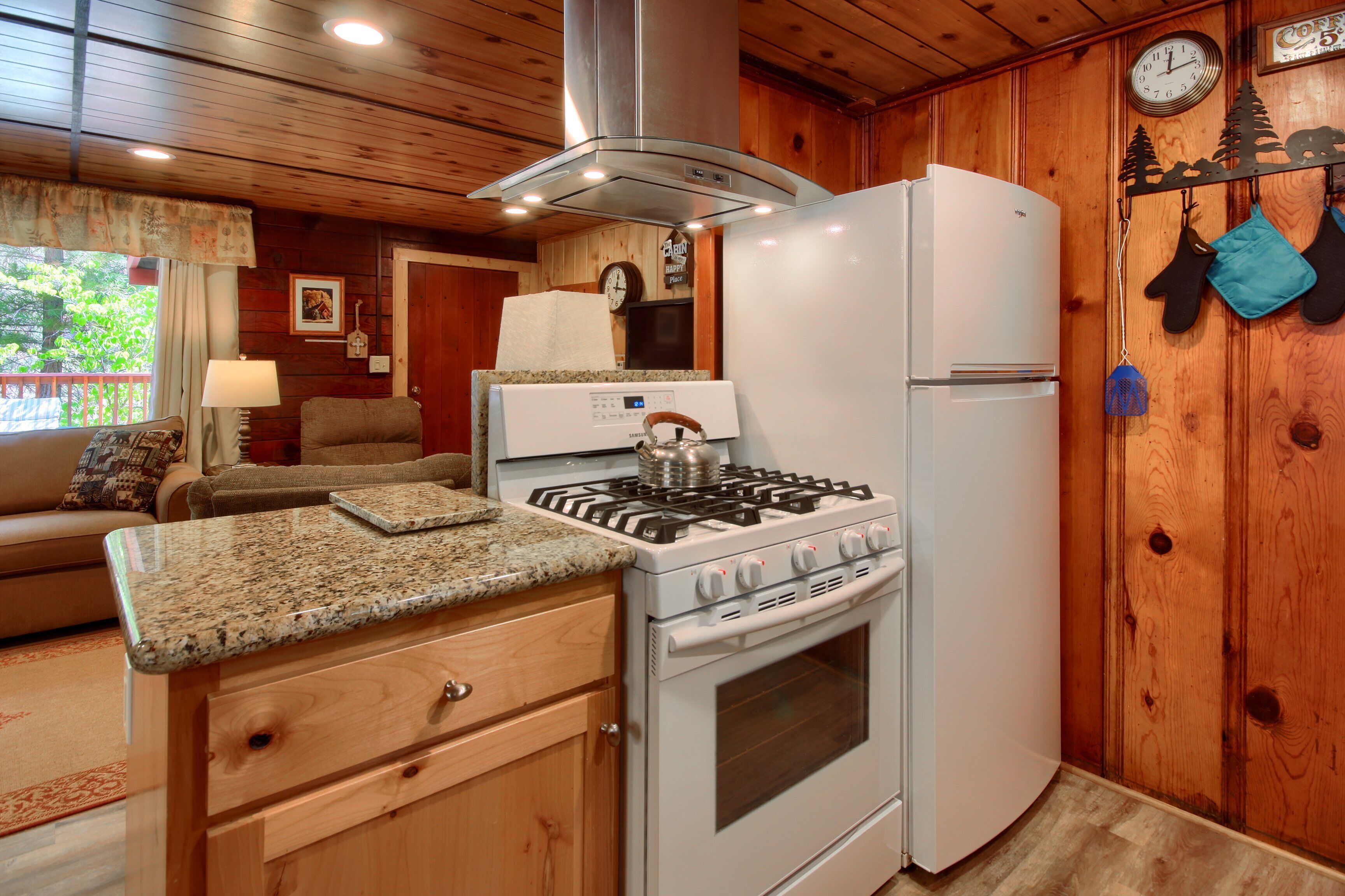 A kitchen with a stove, oven, and refrigerator. There is a clock on the wall and a window with curtains.