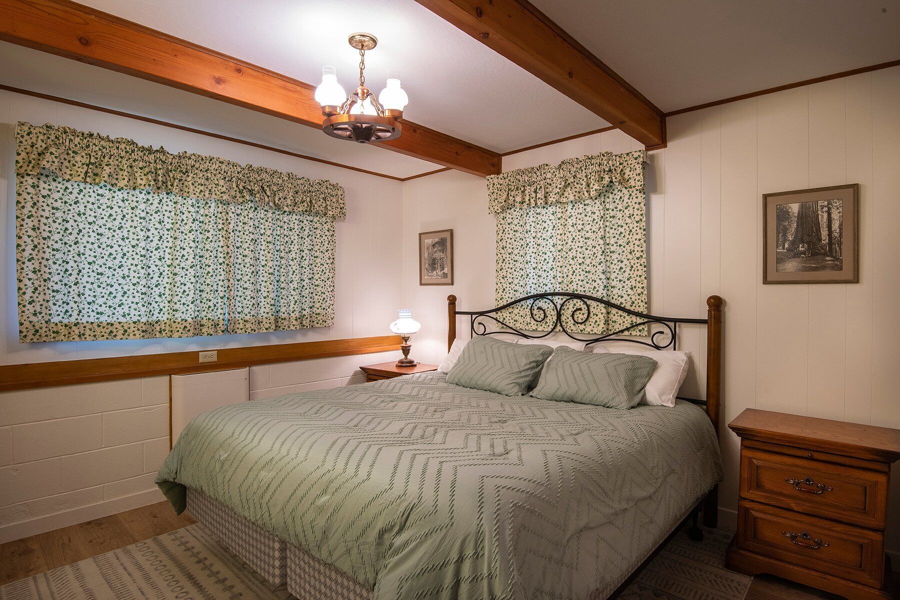 A bedroom with a bed, wooden headboard, a nightstand, a dresser, and a window with floral curtains.