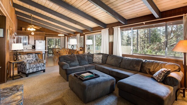 A spacious living room with a large sectional sofa, a small sofa, and a coffee table. There is a dining area with wooden chairs and a table, and a kitchen area with a refrigerator and stove. The room has large windows with curtains, allowing natural light to enter. - Bird's Eye View (Yosemite National Park)