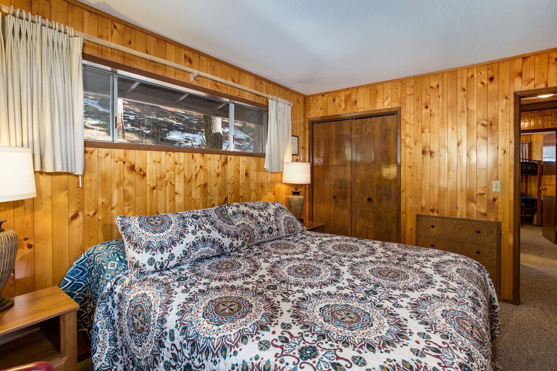 A wooden-paneled room with a bed, a window, a door, and a lamp.