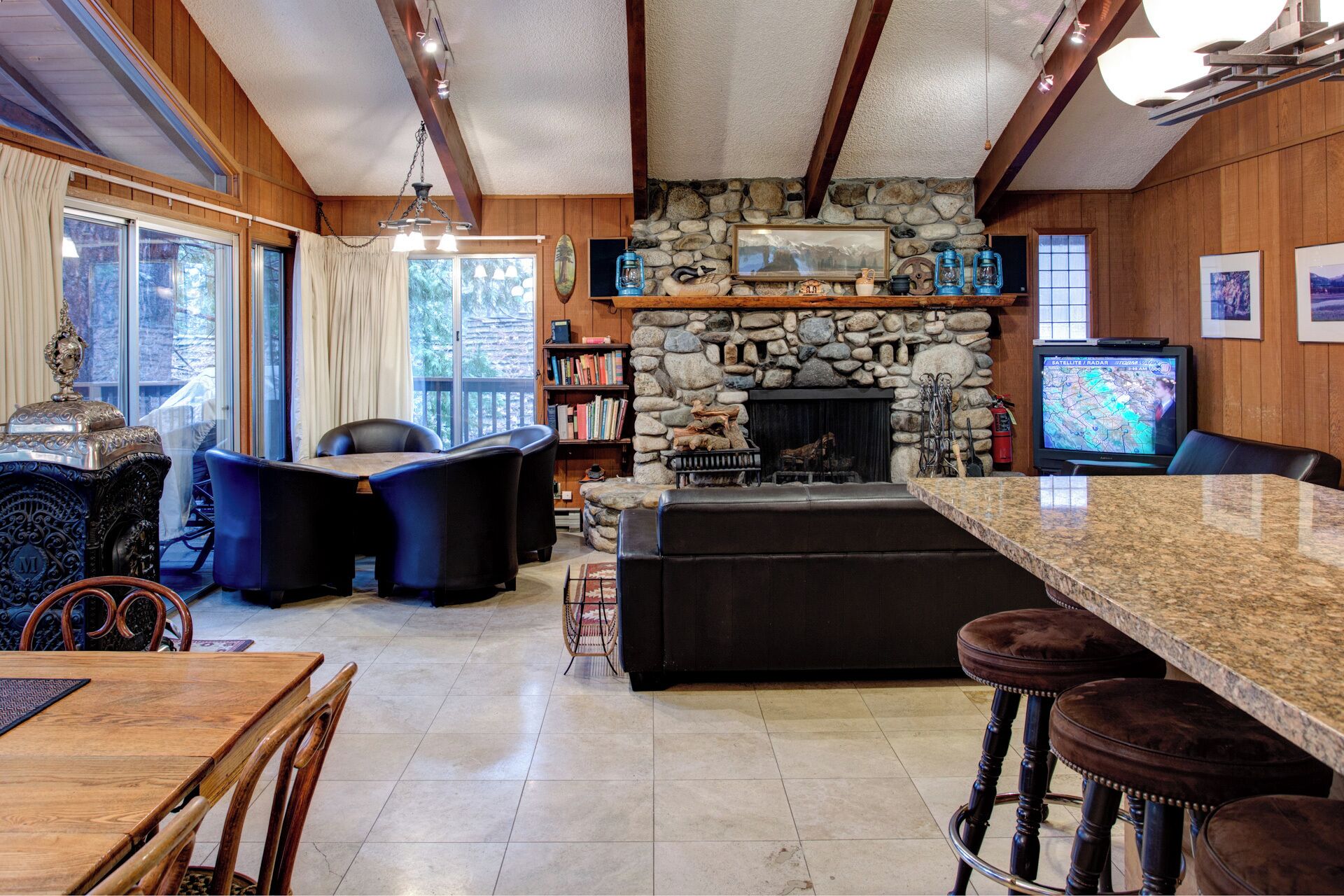 A cozy cabin interior with a stone fireplace, wooden beams, a stone wall, a television, and a dining area with a table and chairs.