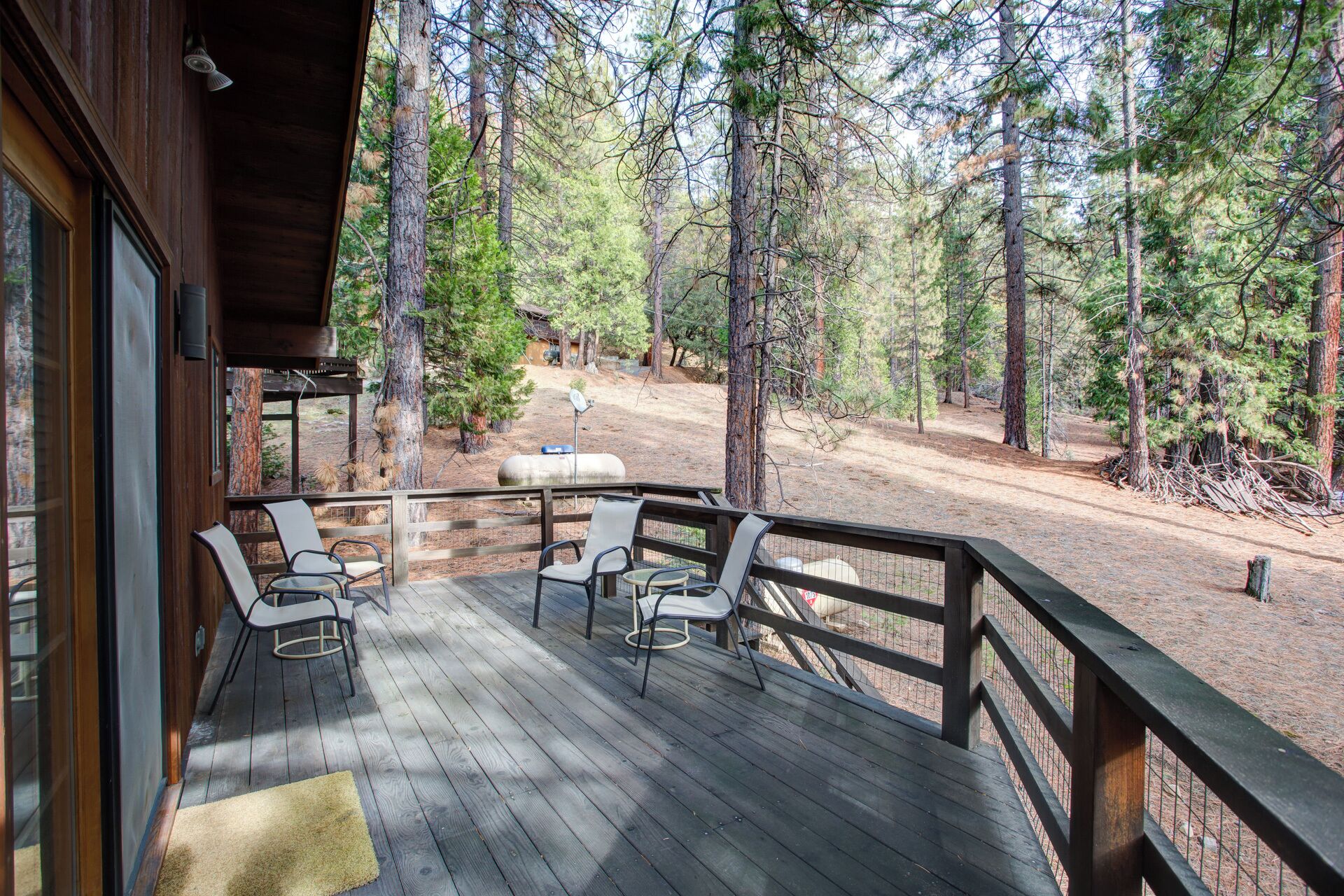 A wooden deck with two chairs and a rug, overlooking a forested area with a dirt path and a small building in the background.