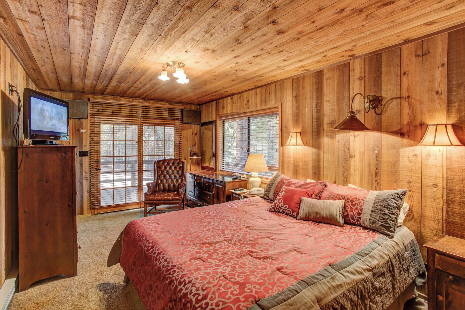 A cozy room with a wooden ceiling, a large bed with patterned bedding, a television on a wooden cabinet, and a desk with a lamp.