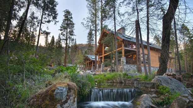 A log cabin with a waterfall in front, surrounded by tall trees. - Rustic Retreat (Yosemite National Park)
