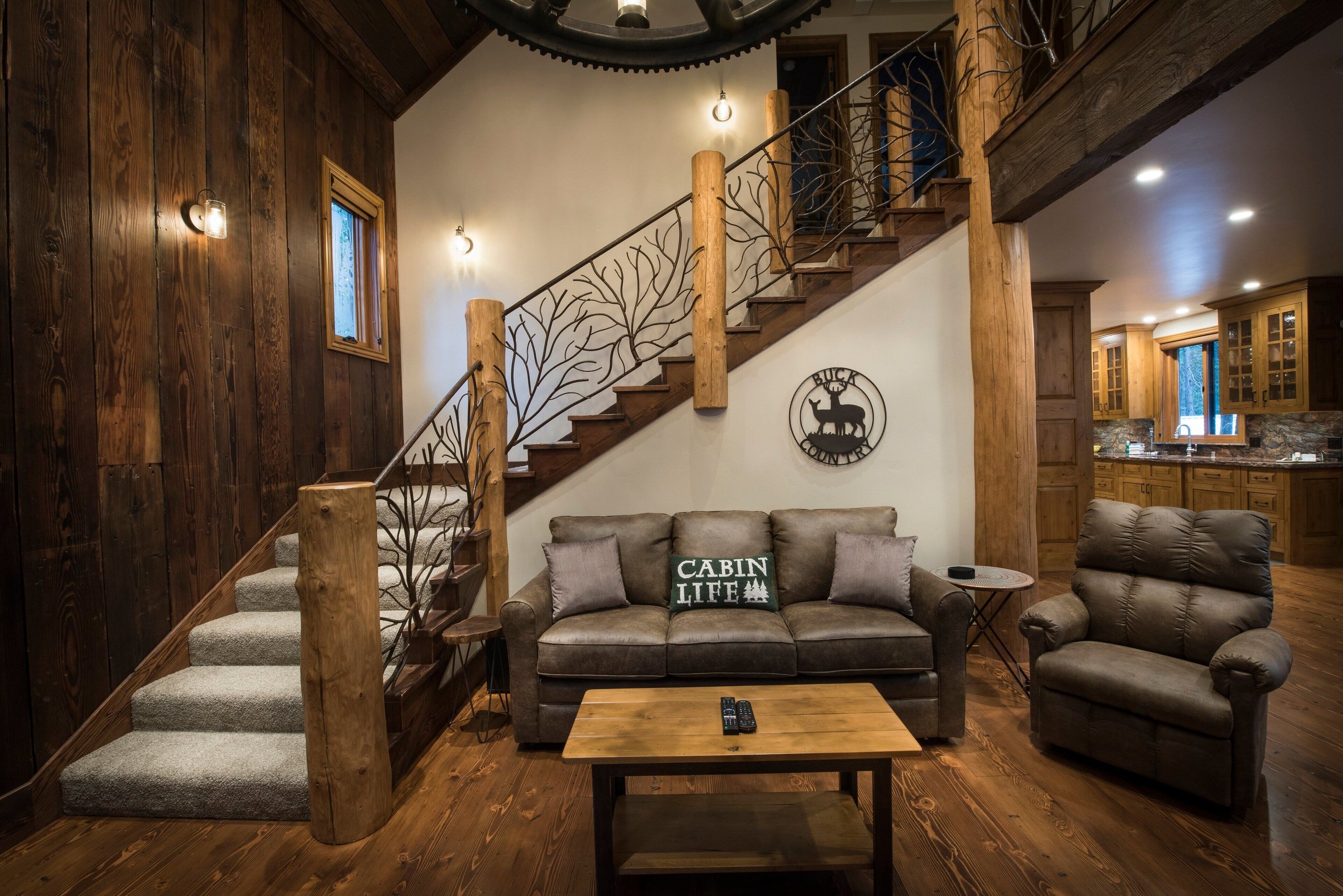 Un intérieur de style cabane avec un escalier en bois, un canapé agrémenté d’un coussin orné de l’inscription « Cabin Life », un fauteuil en cuir et une table basse en bois.