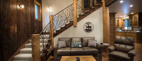 Un intérieur de style cabane avec un escalier en bois, un canapé agrémenté d’un coussin orné de l’inscription « Cabin Life », un fauteuil en cuir et une table basse en bois.
