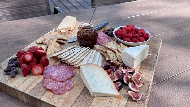 A charcuterie board with various cheeses, fruits, and crackers on a wooden board, set on an outdoor table with chairs. - Heart of the Meadow (Yosemite National Park)