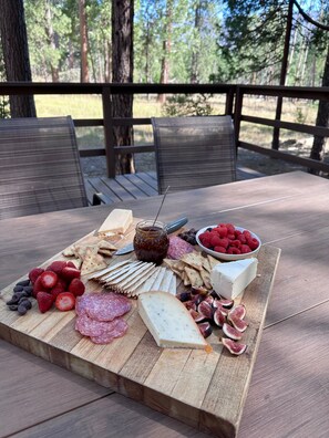 Eine Platte mit verschiedenen Käsesorten, Früchten und Crackern auf einer Holzplatte, platziert auf einem Tisch im Freien mit Stühlen.