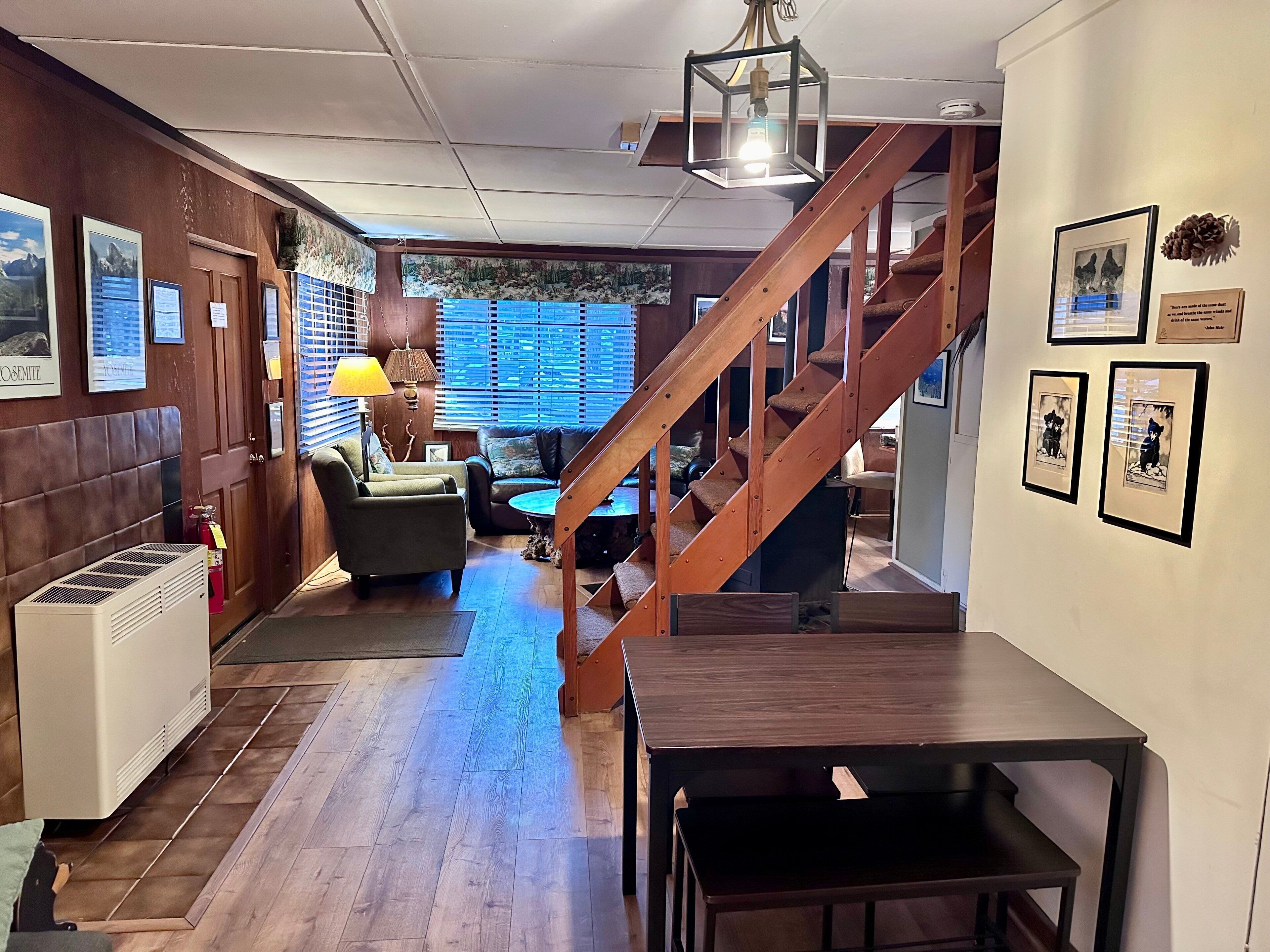 A cozy room with a wooden staircase, a sofa, and a table. There are framed pictures on the wall and a lamp on the table.