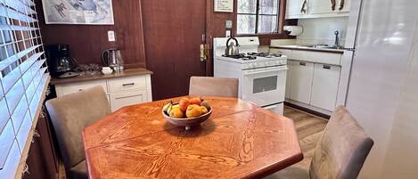 A kitchen with a wooden dining table, chairs, a bowl of fruit, a stove, and a refrigerator.