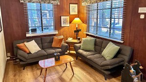 A cozy living room with two grey sofas, a wooden coffee table, and a lamp. Large windows with blinds offer views of the city. - Heart of the Meadow (Yosemite National Park)