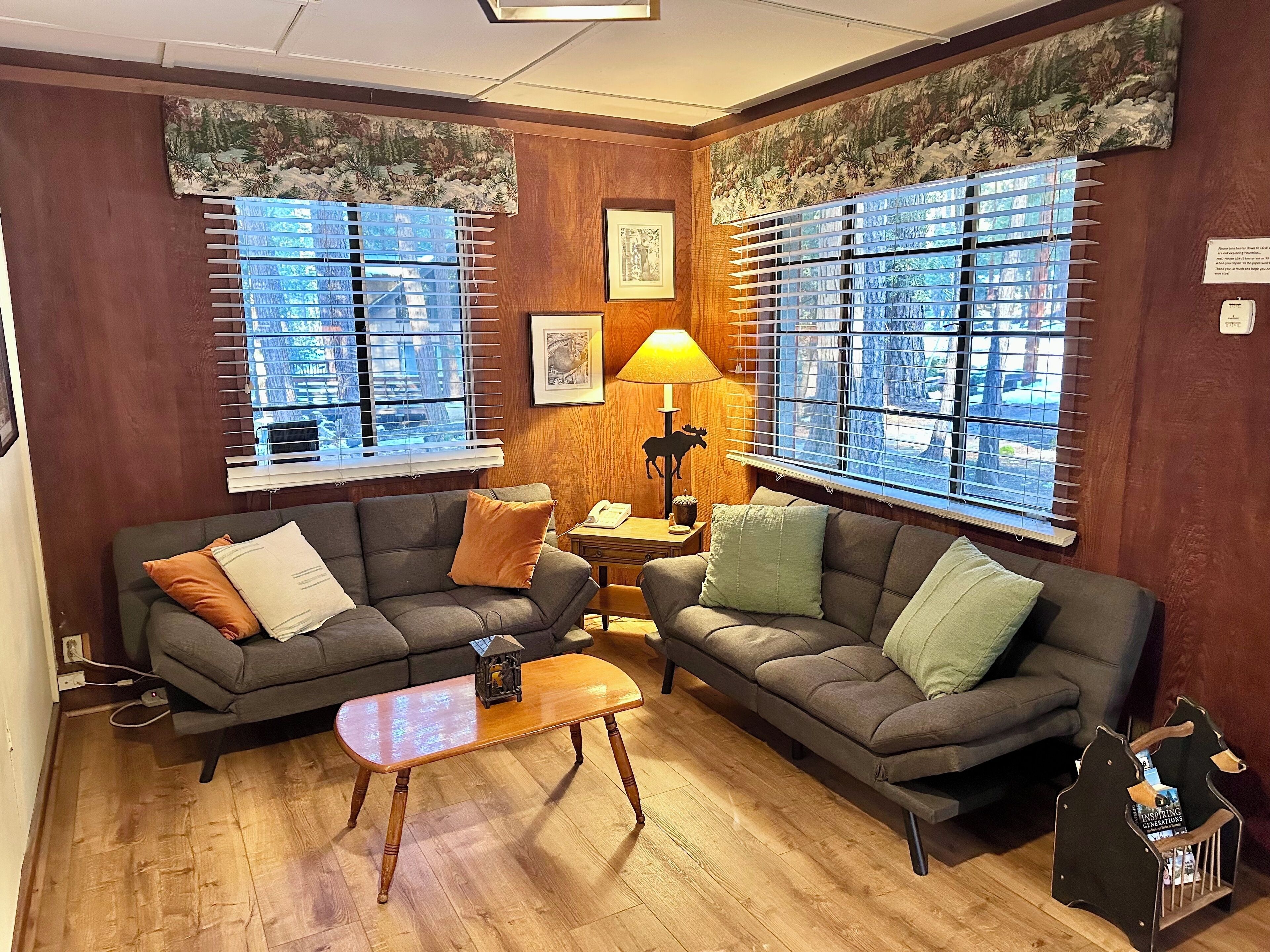 A cozy living room with two grey sofas, a small wooden coffee table, and a lamp. Large windows with blinds provide views of the city.