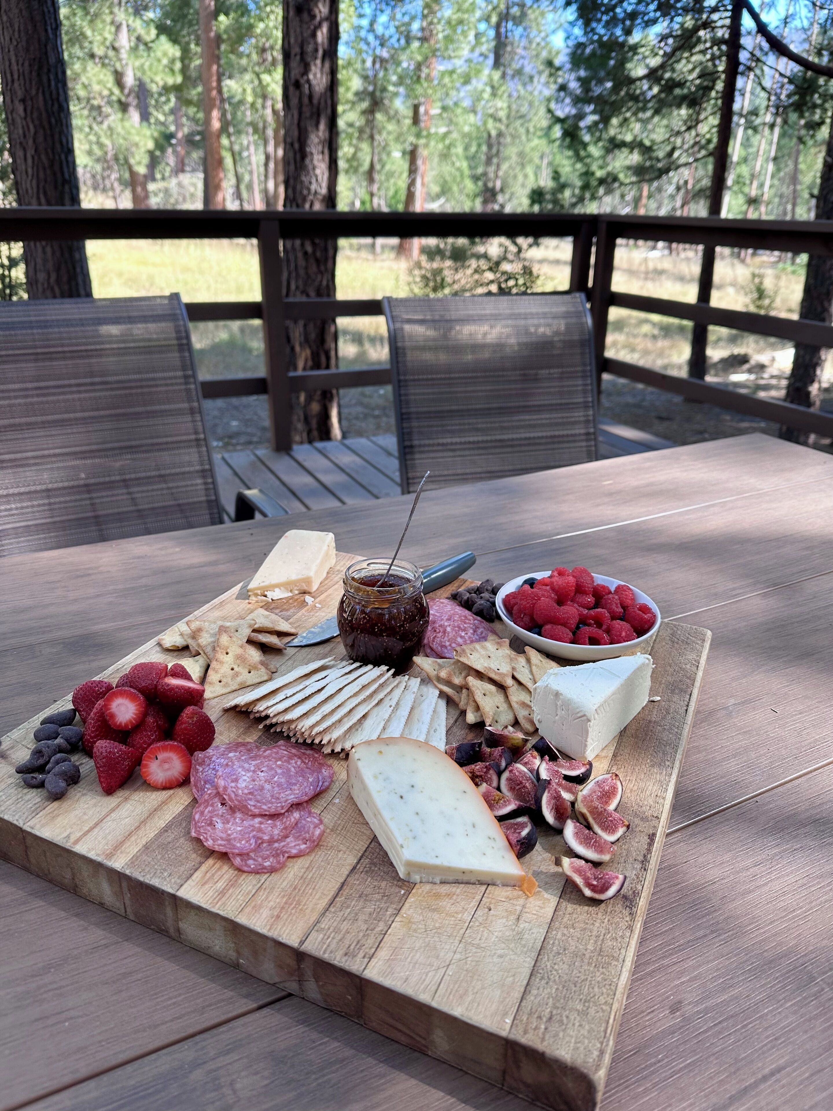 A wooden board with various cheeses, fruits, and crackers on an outdoor table.