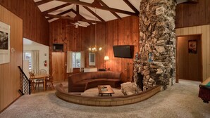 A cozy living room with a stone fireplace, wooden ceiling beams, a television, and a dining area with a table and chairs. - Villarreal Retreat (Yosemite National Park)