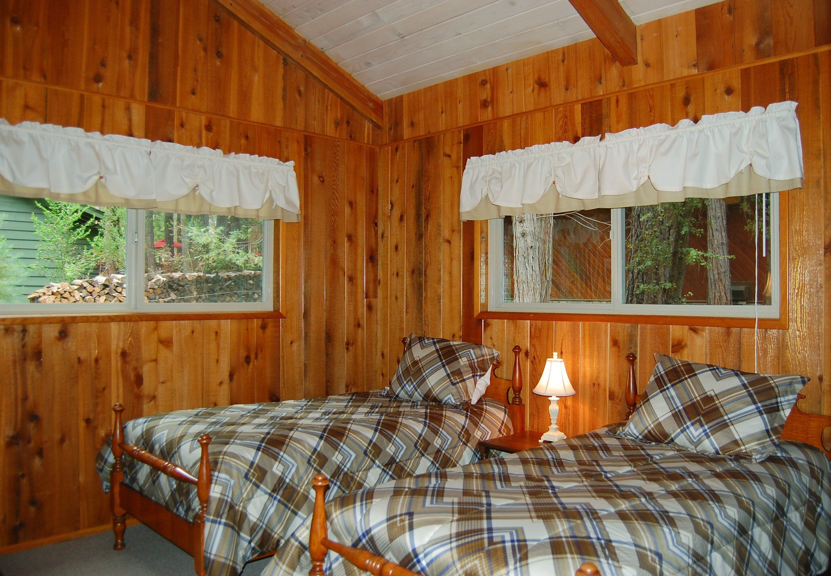 A wooden cabin bedroom with two beds, white curtains, and a nightstand with a lamp.