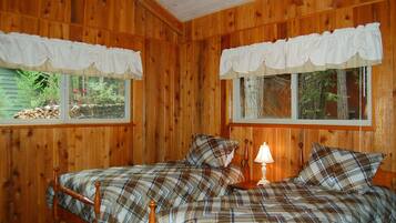 A wooden cabin bedroom with two beds, white curtains, and a nightstand with a lamp.