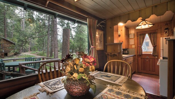 A rustic cabin interior with a dining table set for two, a woven basket with flowers, and a view of the surrounding forest through large windows. - Chipmunk Hollow (Yosemite National Park)