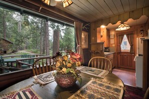 A rustic cabin interior with a dining table set for two, a woven basket with flowers, and a view of the surrounding forest through large windows. - Chipmunk Hollow (Yosemite National Park)