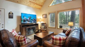 Una sala de estar acogedora con sofás de cuero, un televisor de pantalla plana, chimenea y ventanas amplias con vistas a un bosque.