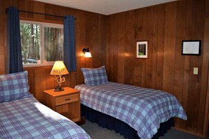 Una habitación con dos camas, una mesita de noche con lámpara, una ventana con cortinas y paredes de madera.