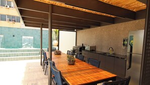 Een moderne eethoek buiten met een houten tafel en stoelen, een keuken met een koelkast en uitzicht op het zwembad.