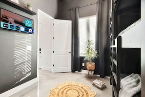 A modern living room with a flat-screen TV displaying streaming services, a white door, a large window with blinds, a woven rug, and a potted plant. - Nashville hot tub & karaoke game room home, sleeps 13, 4 bedrooms (Nashville)